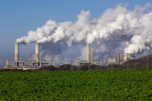 Imagem mostra uma indústria emitindo grande quantidade de fumaça no céu, provavelmente um processo de emissão de gases de efeito estufa.